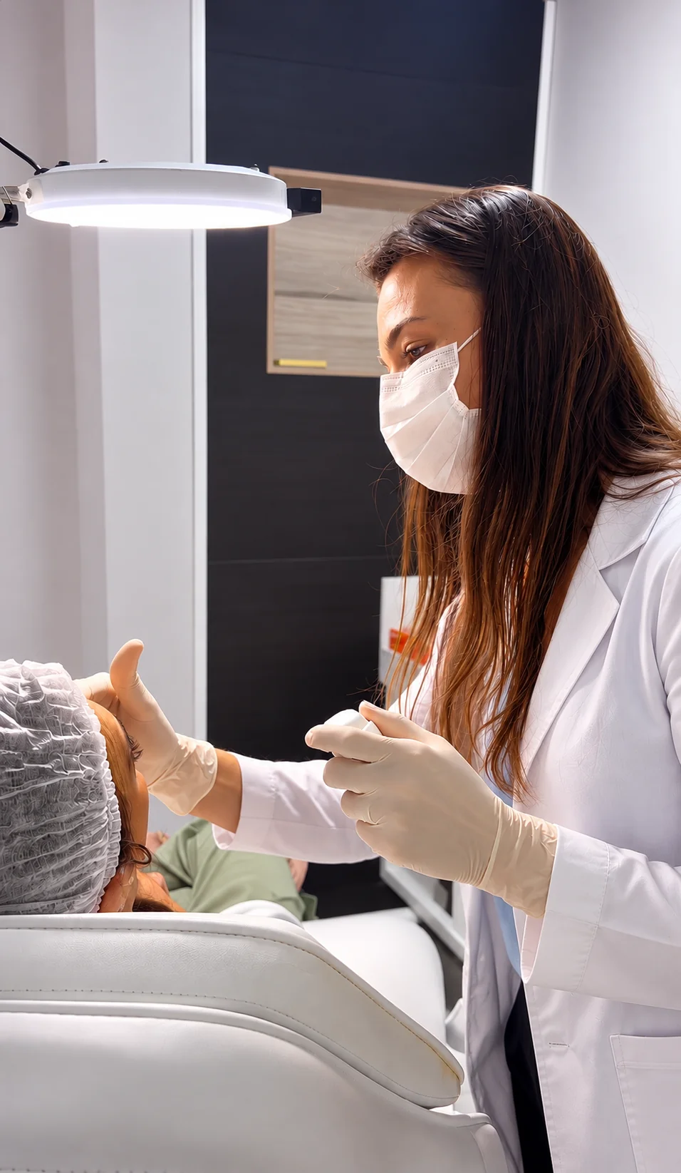 Dra. Julha Sbardeloto realizando um procedimento facial no consultório, sob luz de exame